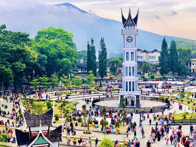 Menara Jam Gadang utuh di tengah pelataran yang ramai pejalan kaki, dengan pepohonan dan gunung di latar.