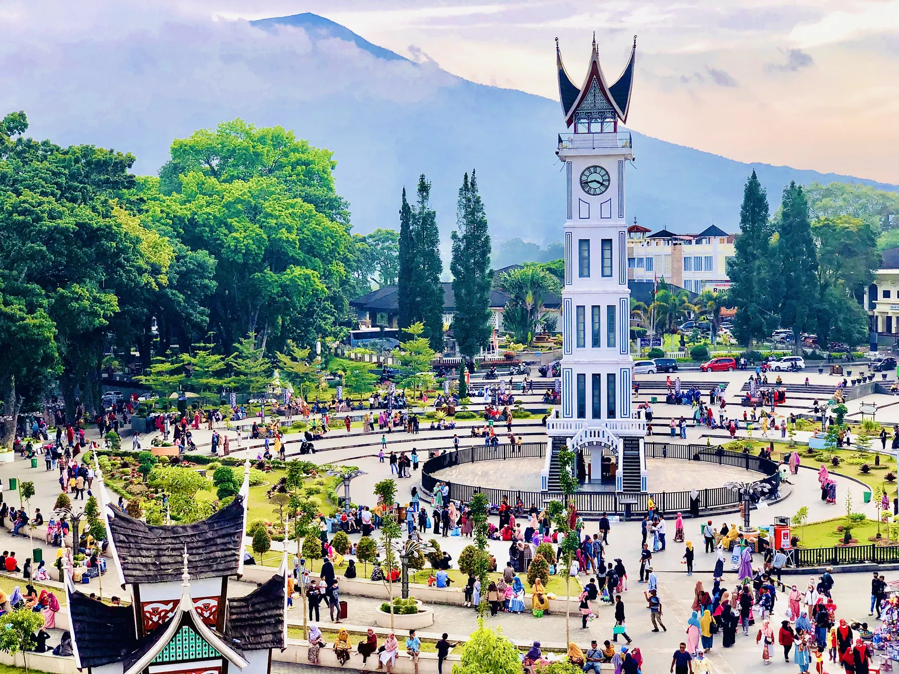 Menara Jam Gadang utuh di tengah pelataran yang ramai pejalan kaki, dengan pepohonan dan gunung di latar.