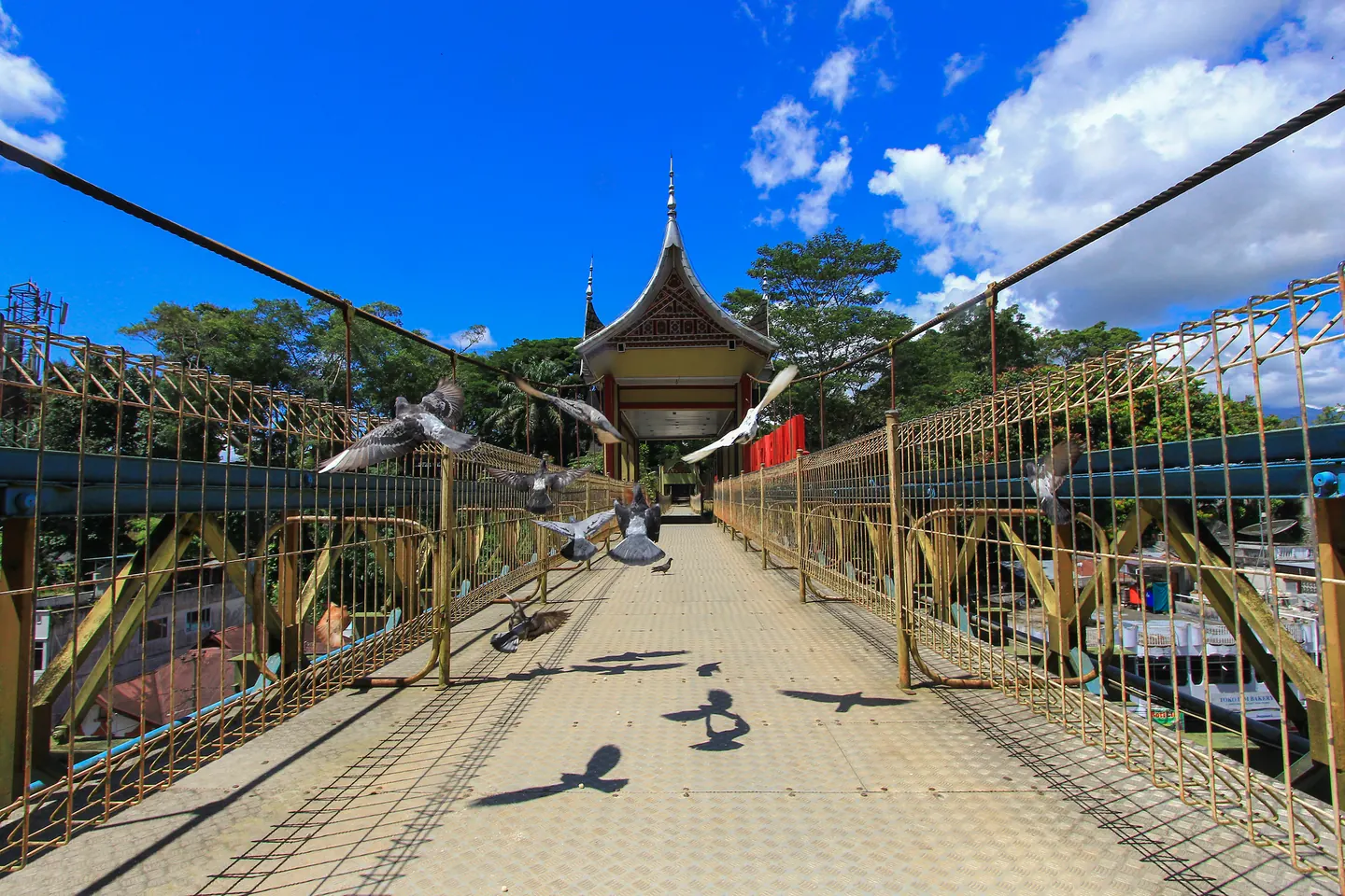 Jalur pejalan kaki Jembatan Limpapeh dengan pagar di kedua sisi, paviliun beratap Minangkabau, dan burung yang sedang terbang.
